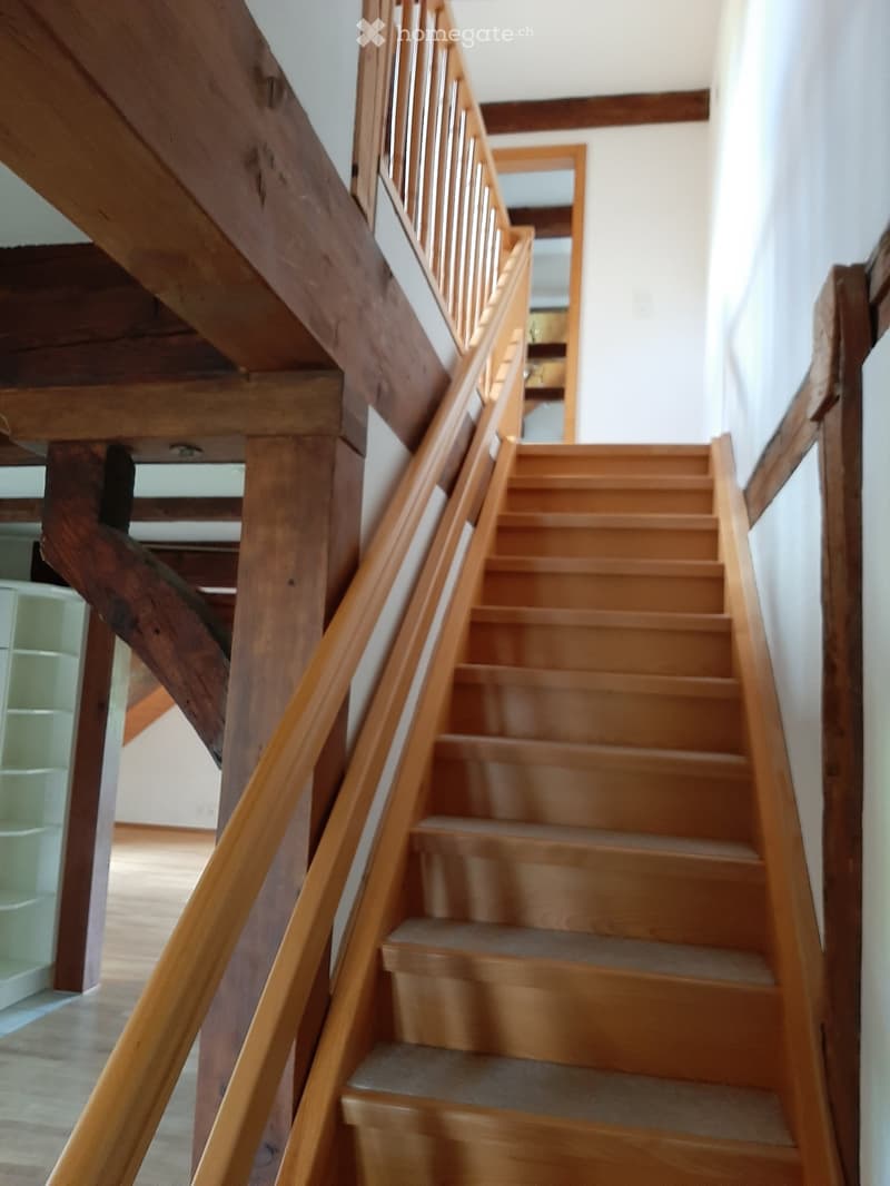 Wooden staircase with a handrail and wooden steps, leading upwards. The walls are white, and the ceiling has exposed wooden beams.