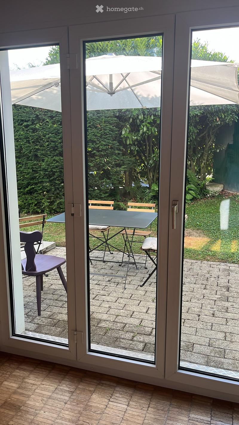 View through large glass doors to a patio area with a table, chairs, and an umbrella, surrounded by greenery.