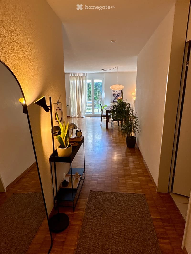 A well-lit hallway with wooden flooring, a small side table with a potted plant, a floor lamp, and a view into a dining area with a table and chairs.