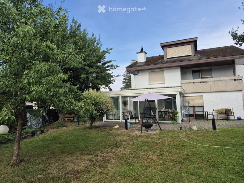 Lawn with trees, patio area with outdoor furniture, umbrella, and a modern house with large windows and a balcony.