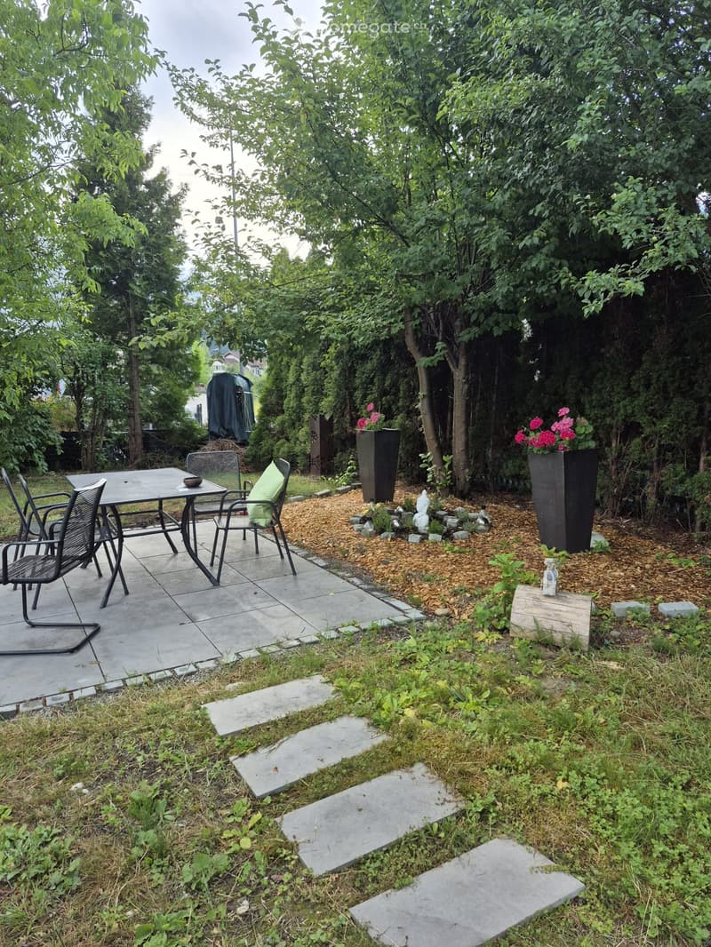 Garden area with a patio, table, chairs, and flower pots surrounded by trees and greenery.