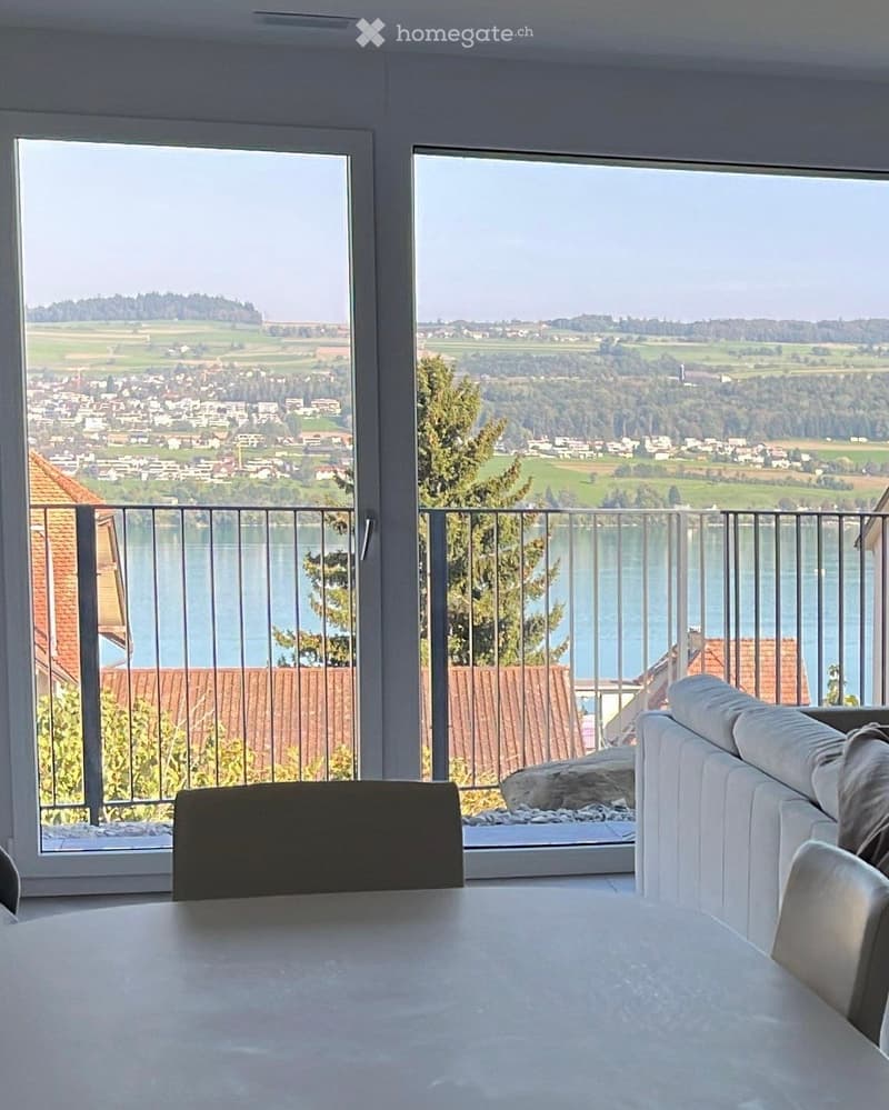 Bright room with large windows offering a scenic view of a lake and surrounding landscape. Features a white sofa, a round table, and a chair.