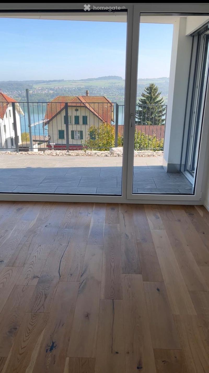 Empty room with light wooden flooring and large sliding glass doors leading to a balcony with a scenic view of a lake and surrounding landscape.