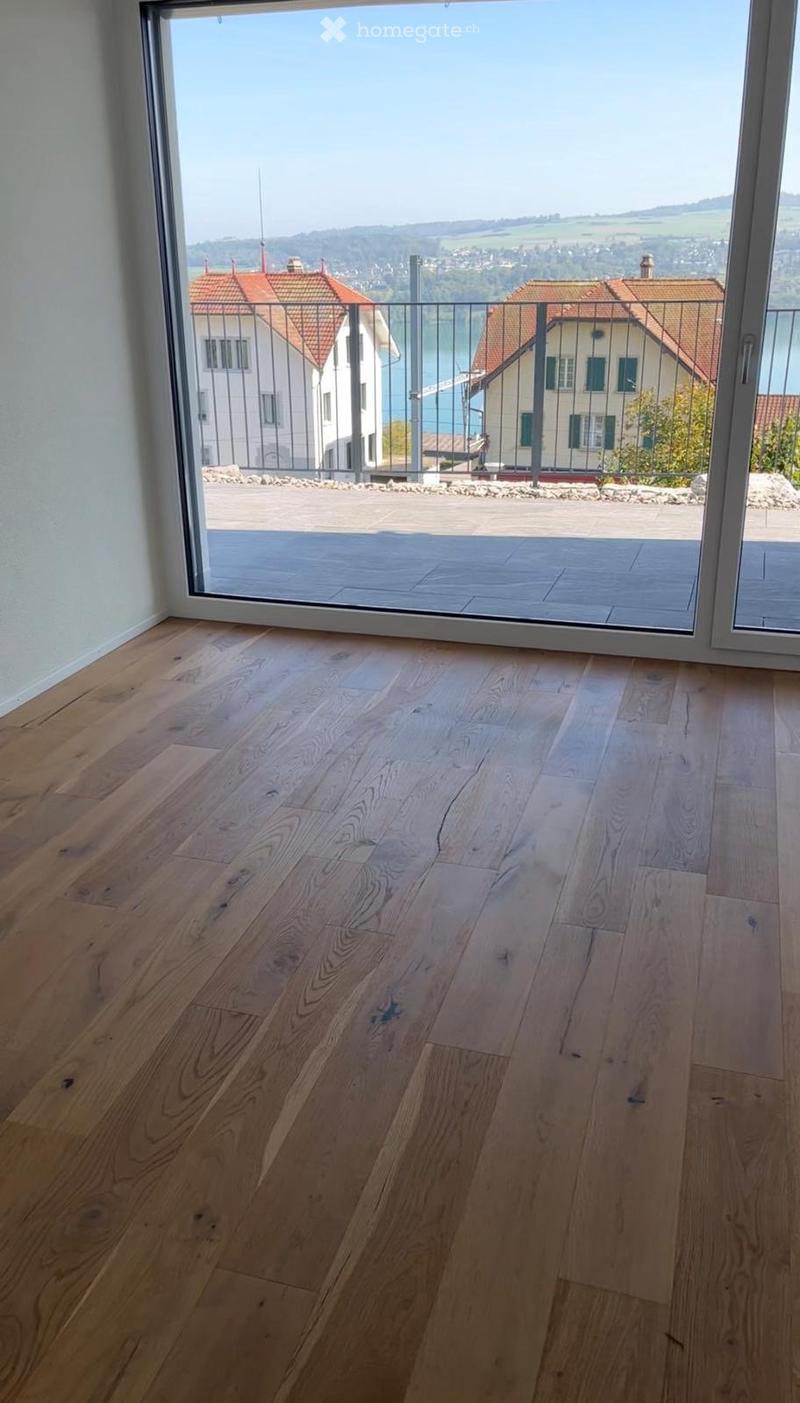 Empty room with light wooden flooring and large glass sliding doors leading to a balcony with a scenic view of a town and lake.