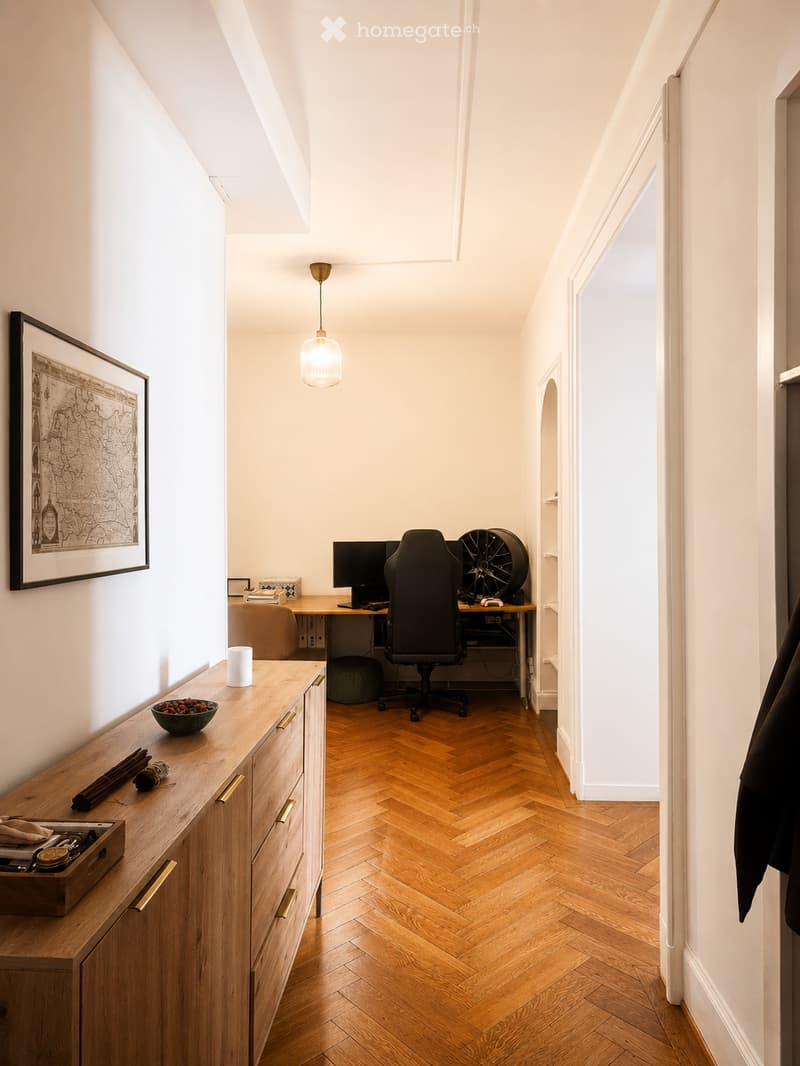 A modern, well-lit room with herringbone wooden flooring, a desk with a computer and chair, a hanging lamp, a wooden cabinet with drawers, and decorative items on the cabinet.