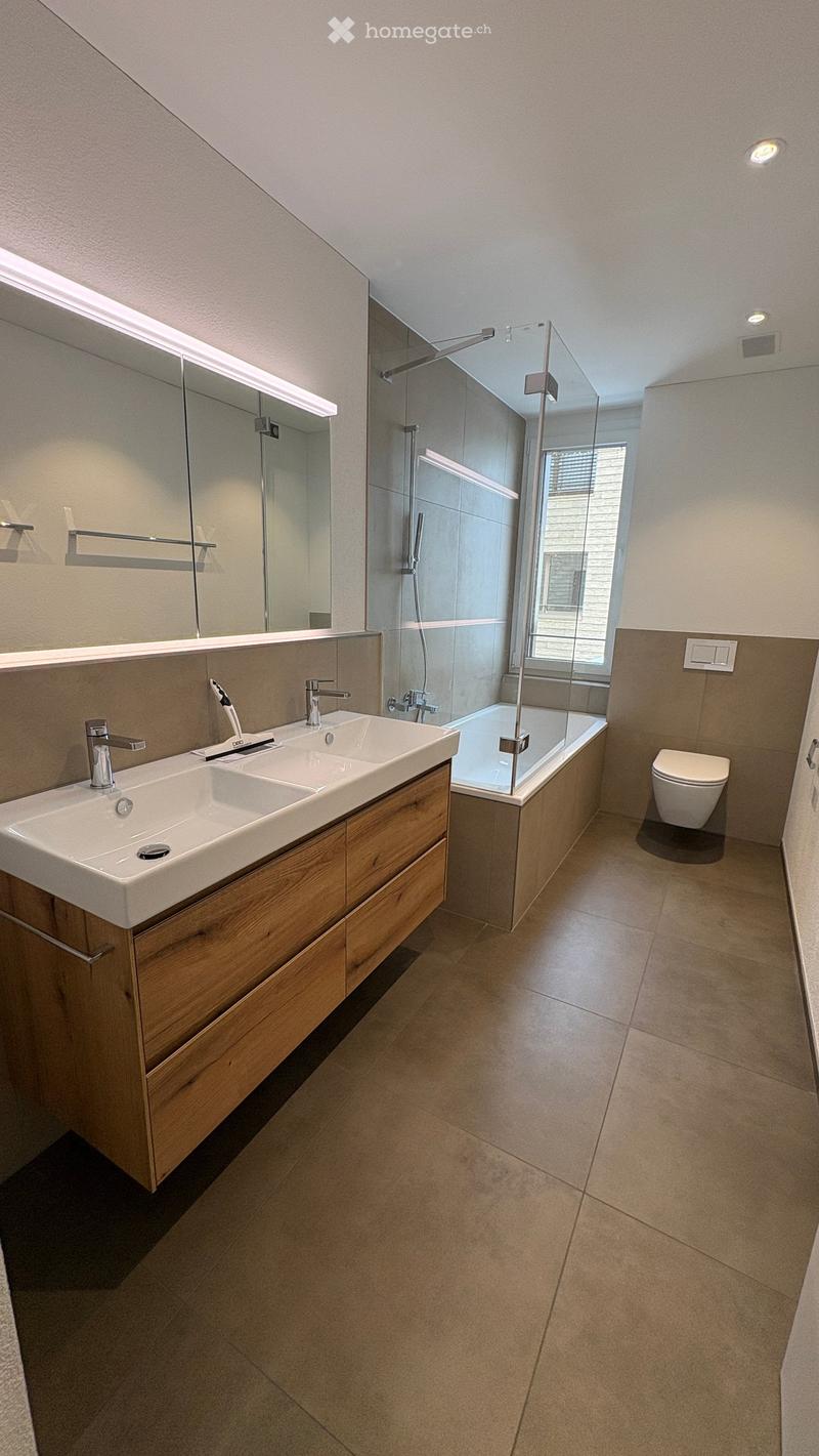 Modern bathroom with double sink, wooden cabinet, large mirror, bathtub with glass shower enclosure, and toilet.