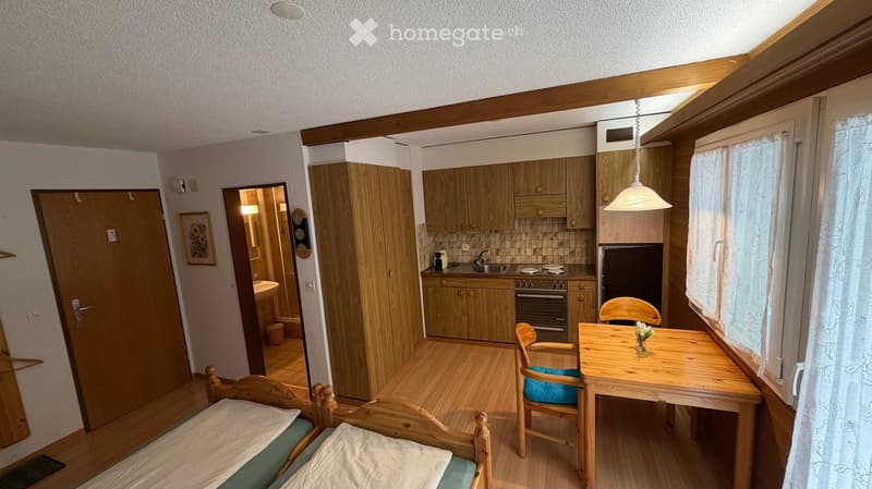 A compact, furnished room with a bed, a small table and chairs, a kitchen area with cabinets, a fridge, an oven, and a sink. The room has wooden flooring and a slanted ceiling with a skylight.
