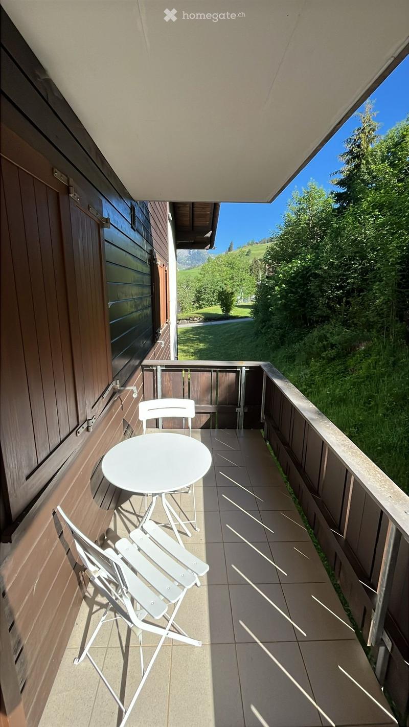 Balcony with a white table and two white chairs, overlooking a green landscape with trees and mountains.