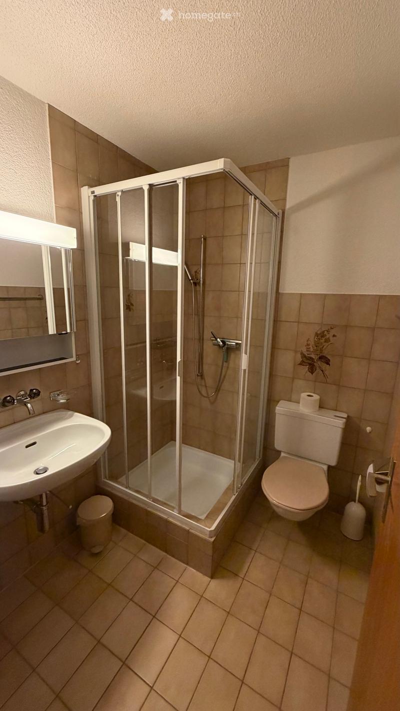 Modern bathroom with a glass-enclosed shower, a white sink, a toilet, and a mirror. The walls are tiled in beige, and the floor is covered with light-colored tiles.
