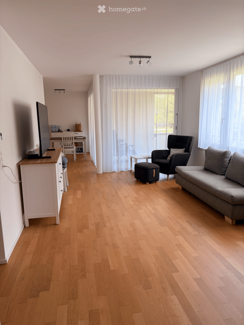 Bright living room with hardwood flooring, large windows with sheer curtains, grey sofa, black ottoman, coffee table, TV stand, and dining area with table and chairs.