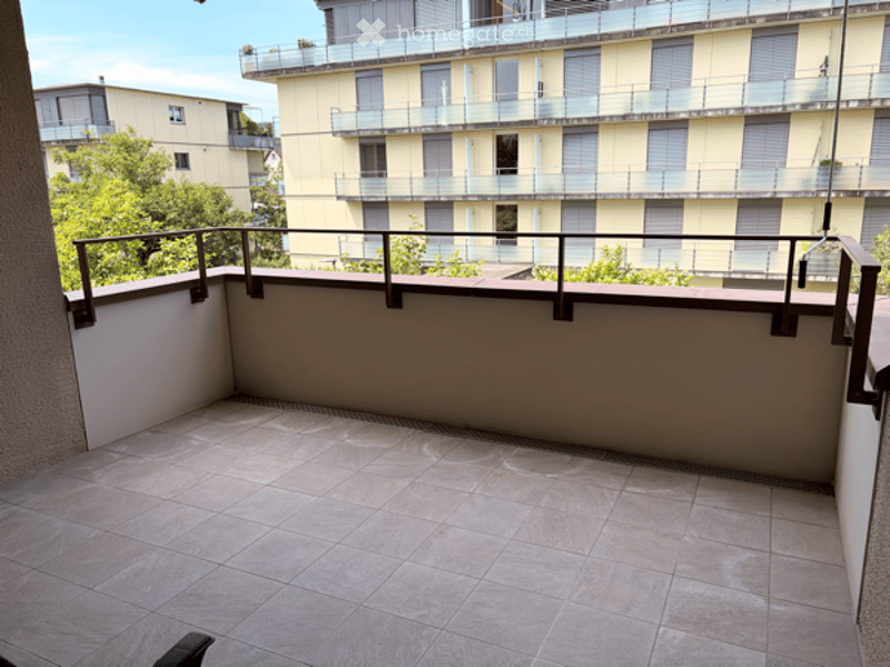 Balcony with tiled floor, metal railing, overlooking a multi-story building with balconies and greenery.