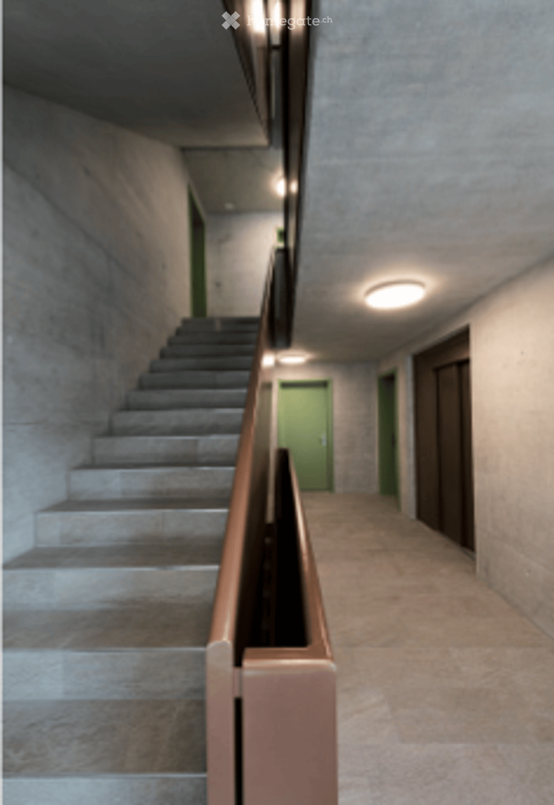 Modern hallway with concrete walls, tiled floor, staircase leading upwards, and green doors visible at the end.