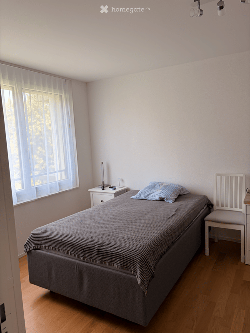 A simple, unfurnished bedroom with a single bed, nightstand, and chair. The room features light-colored walls, wooden flooring, and a window with sheer curtains.