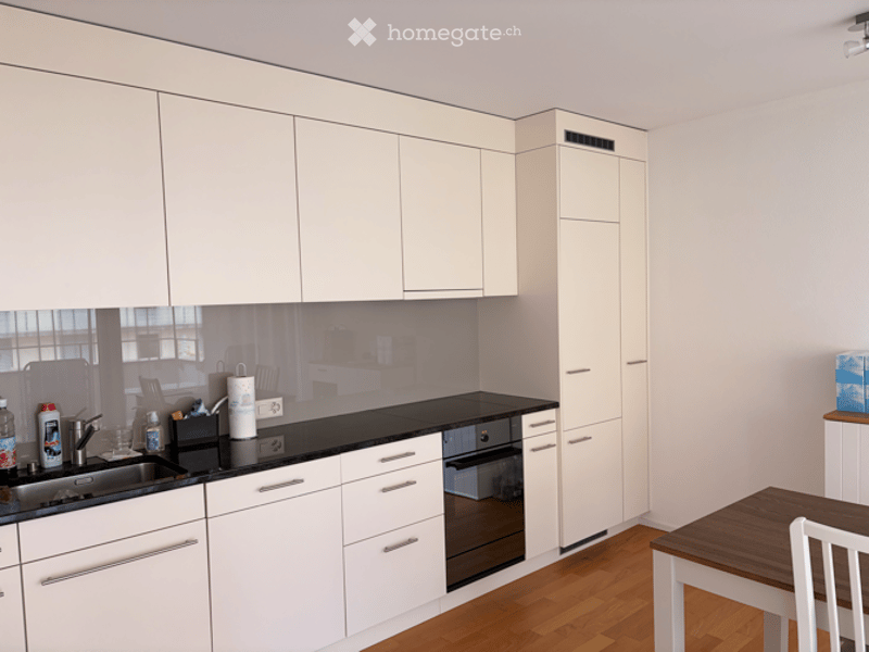 Modern kitchen with white cabinets, black countertops, and a wooden floor. Features a sink, oven, and a small dining area with a table and chair.
