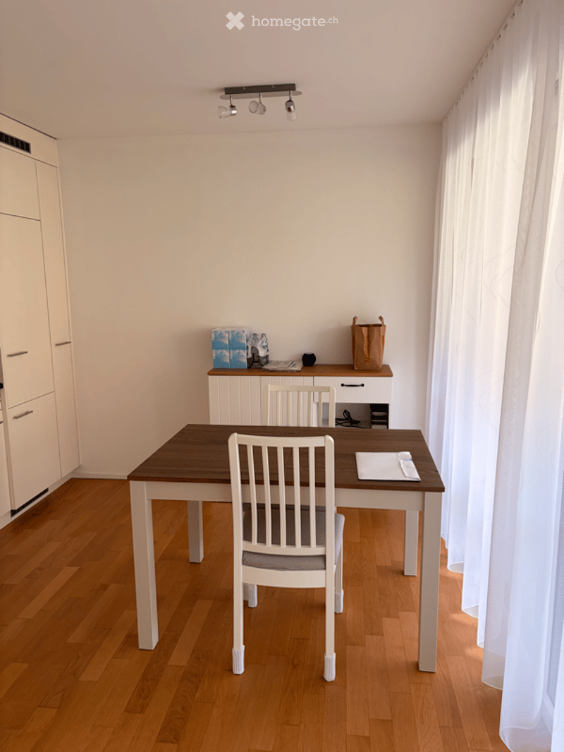 A modern dining area with a wooden table, white chairs, a cabinet, and a laptop on top. The room features wooden flooring, white walls, and large windows with white curtains.
