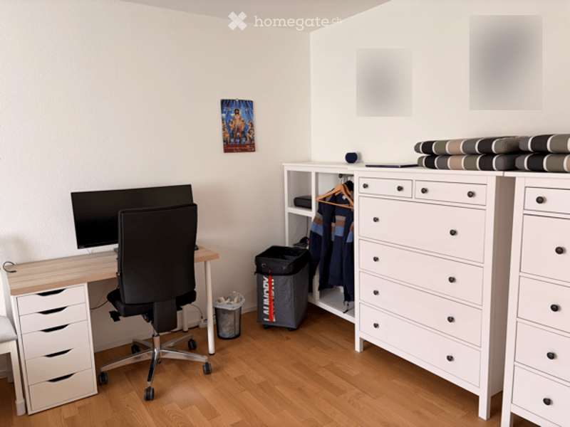 A tidy, modern room with a wooden floor, white walls, and a desk setup featuring a computer monitor, chair, and drawer unit. A wardrobe with folded clothes and a storage box is visible, along with a picture frame on the wall.