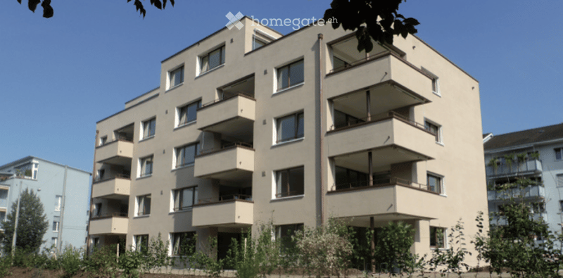Multi-story residential building with multiple balconies and windows, surrounded by greenery.
