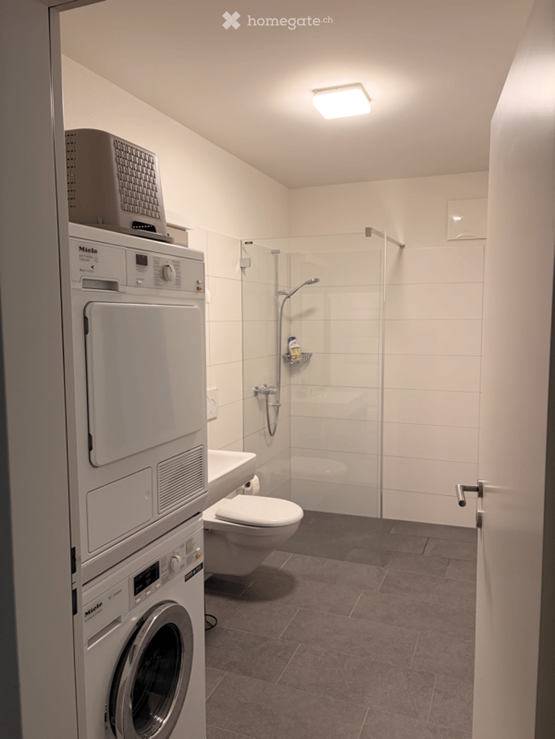 Modern bathroom with a toilet, glass-enclosed shower, and stacked washing machine and dryer unit.