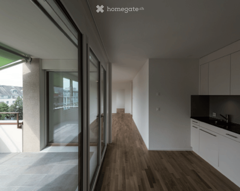 Bright, unfurnished room with wooden flooring, large glass doors leading to a balcony, and modern white cabinetry.