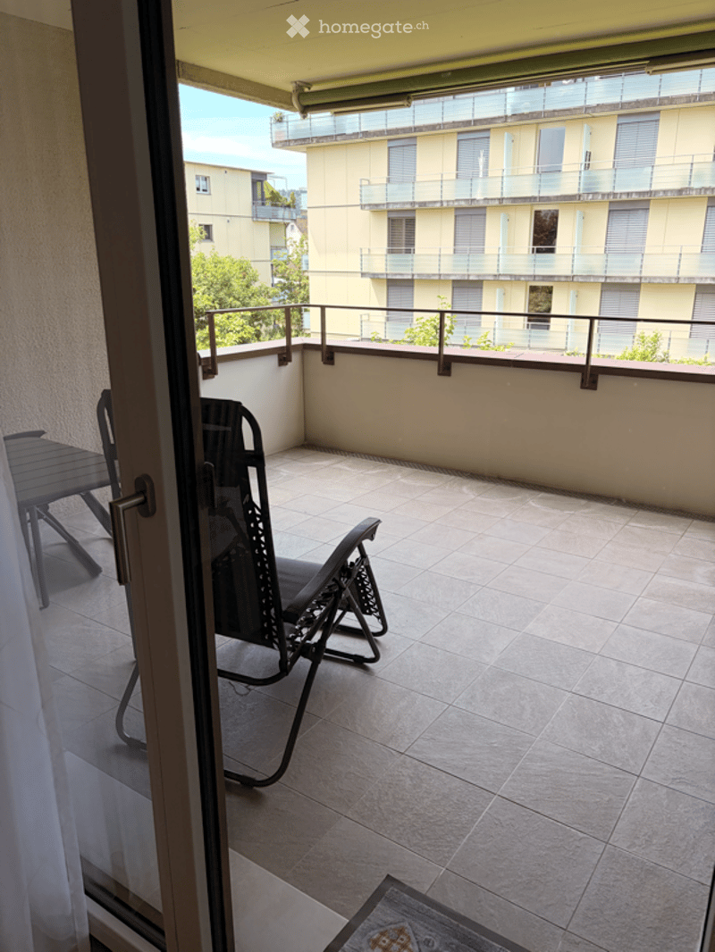 Balcony with tiled floor, black folding chair, small table, and a view of neighboring buildings and greenery.