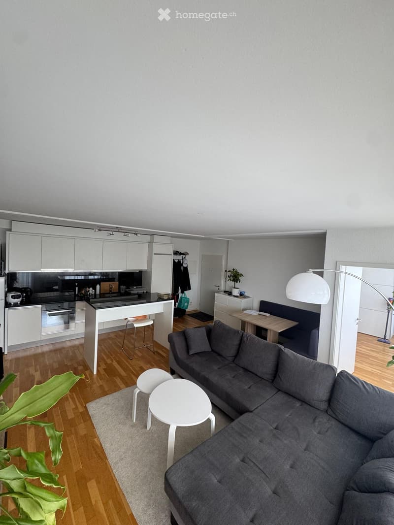 Modern open-plan living and dining area with a grey sectional sofa, white coffee table, white stools, a kitchen island with bar stools, and a dining table with chairs. The space features light grey walls, wooden flooring, and contemporary furniture.