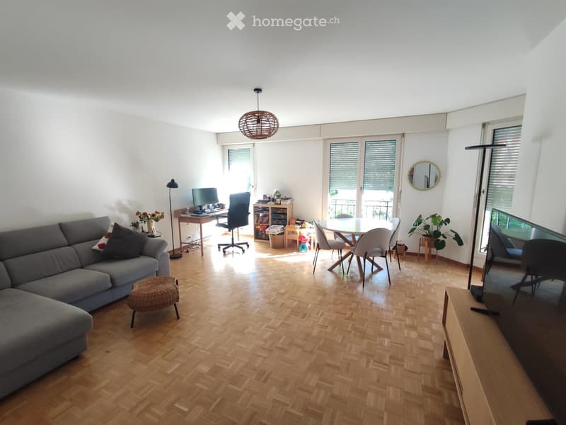 Bright living room with wooden flooring, large windows, and a balcony. Features a grey sofa, a round dining table with chairs, a TV screen, and a desk with a computer.