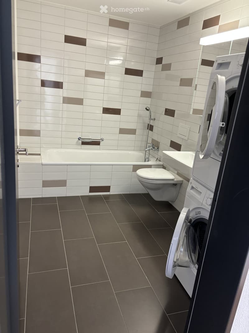Modern bathroom with white and brown tiled walls, a bathtub, toilet, sink, and a washing machine in the corner.