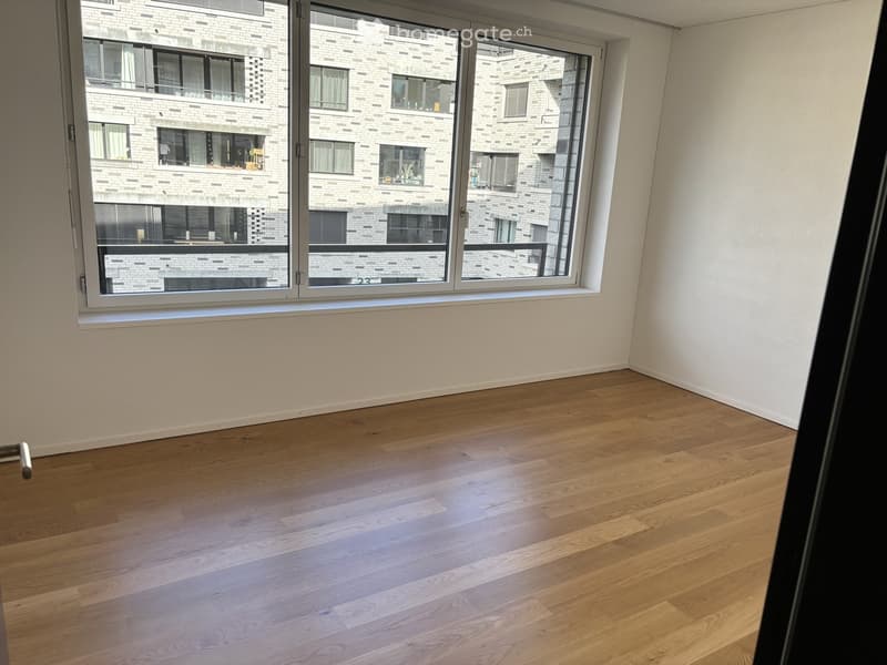 Empty room with light wooden flooring and large windows showing a brick building exterior.