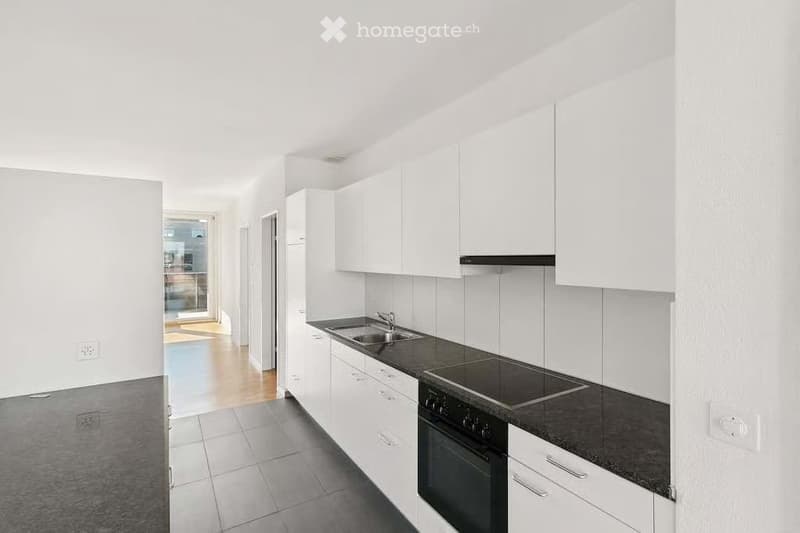 Modern kitchen with white cabinets, black countertops, and stainless steel appliances including a sink, stove, and oven. The space features tiled flooring and a view to an adjacent room through an open doorway.