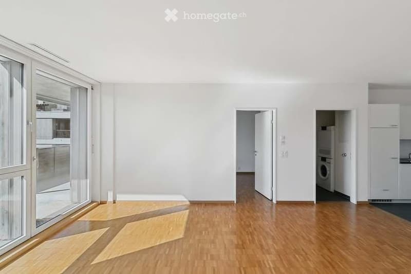 Empty room with wooden flooring, white walls, large windows, and open doors leading to other areas.