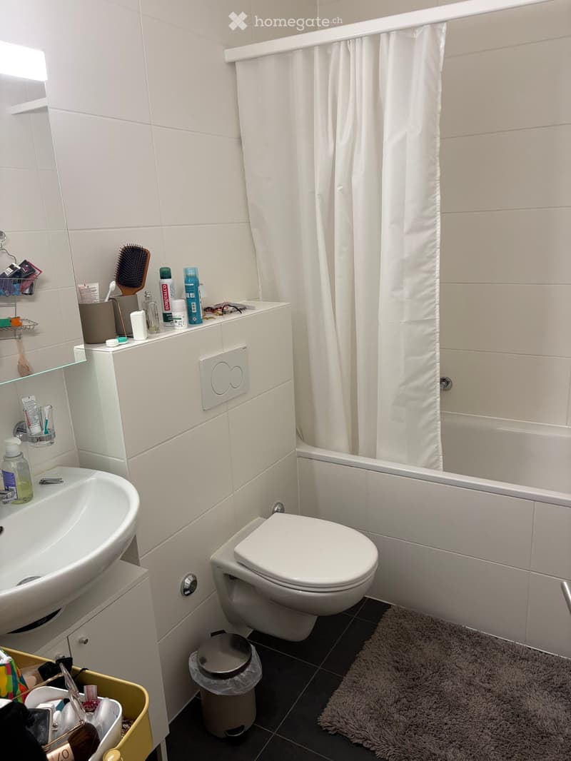 Modern bathroom with white tiles, a sink, toilet, bathtub with a shower curtain, and various toiletries on the counter.