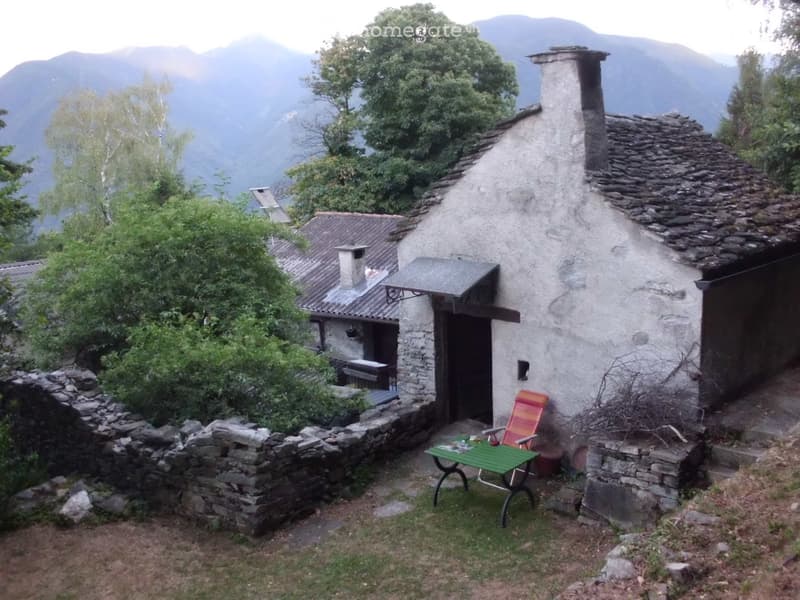A rustic stone cottage with a small outdoor seating area featuring a green table and colorful chair, surrounded by greenery and mountains.