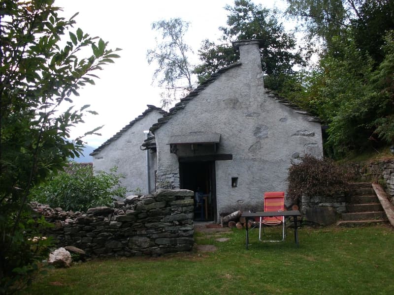 A small, rustic stone cottage with a chimney, surrounded by a grassy area with a stone wall, a table, and a chair.