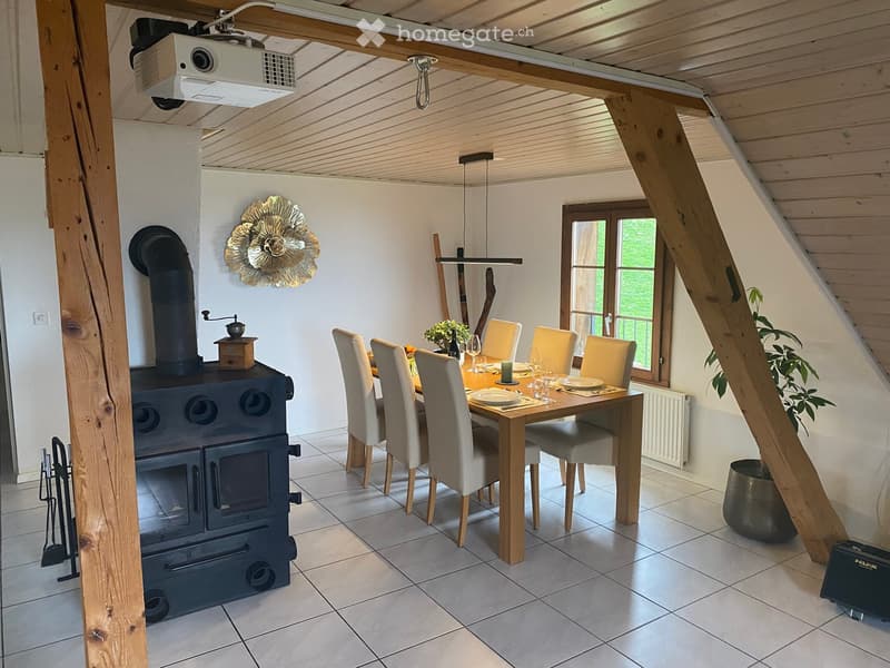 Cozy dining area with a wooden table, six chairs, a black wood-burning stove, and a projector mounted on the ceiling. The room features exposed wooden beams, large windows, and decorative elements including a wall-mounted floral design and a coffee grinde