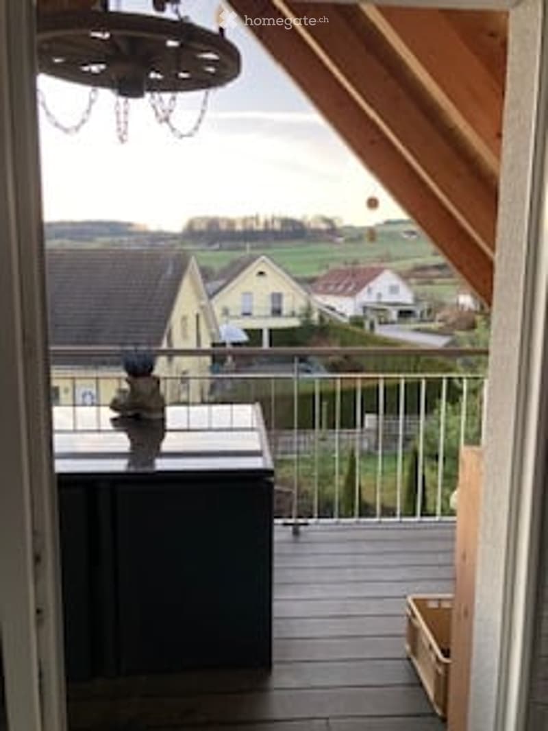 Balcony with wooden flooring, metal railing, small table, and a view of the countryside with houses in the background.
