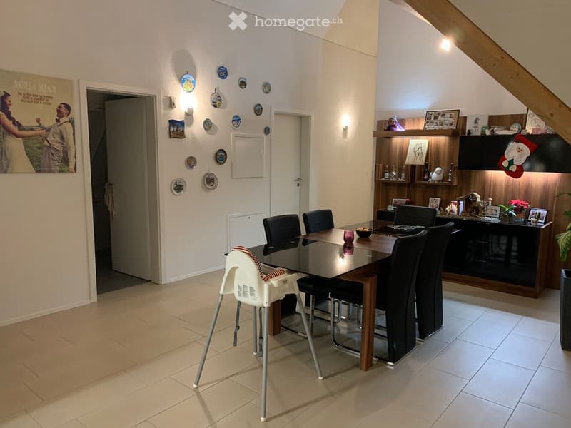 Dining area with a glass dining table, six chairs, a baby chair, and a kitchen counter with various items including a cup, bowl, and framed pictures. The room features a mix of wooden and tiled flooring, and decorative plates on the wall.
