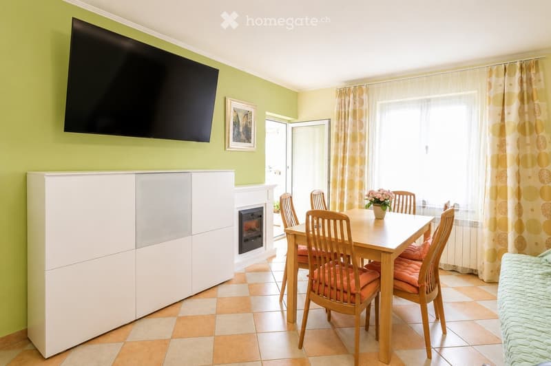 Bright living room with green walls, a large flat-screen TV, a fireplace, a dining table with six chairs, and a vase of flowers on the table.
