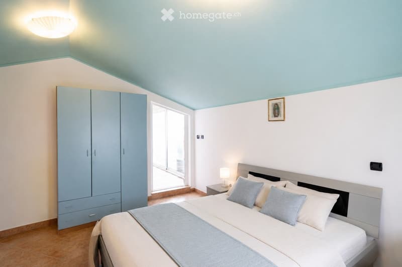 Modern bedroom with a double bed, nightstand, wardrobe, and a picture frame on the wall. The room features light blue walls and a wooden floor.