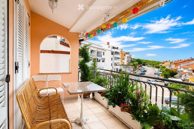 Balcony with a marble table, wicker chairs, and flower pots, overlooking a town with parked cars and buildings.