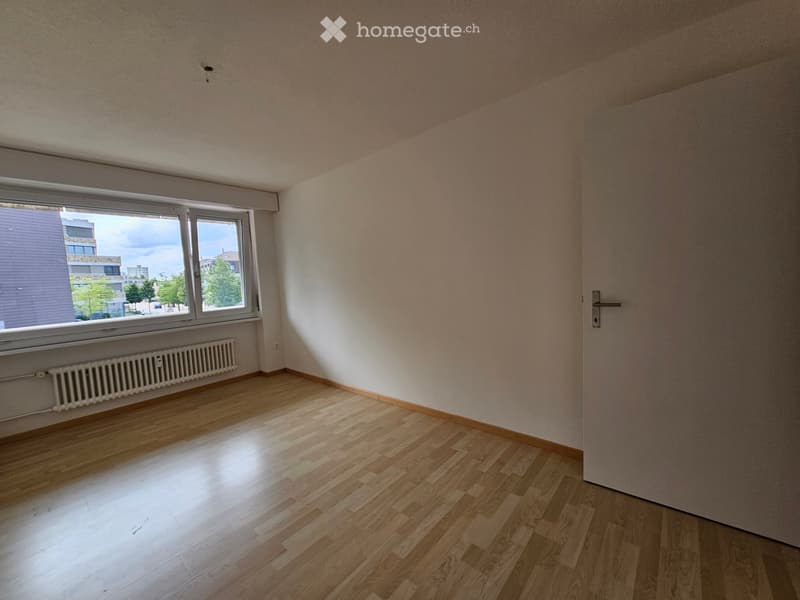 Empty room with wooden flooring, white walls, large window, radiator, and a closed door.