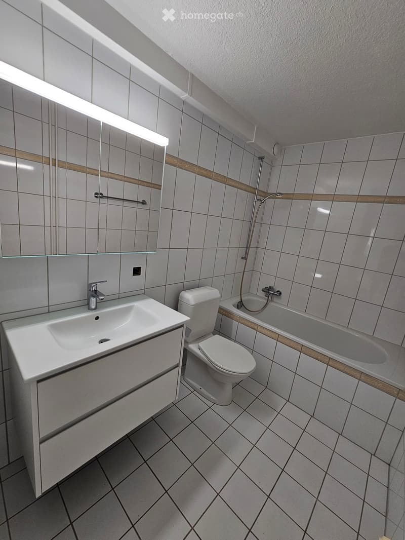 Modern bathroom with white tiles, a sink, toilet, bathtub, and a large mirror with built-in lighting.
