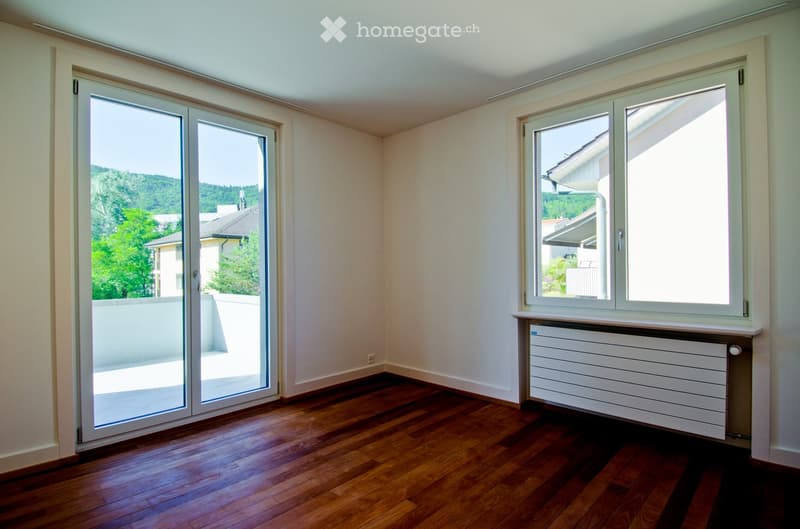 Empty room with wooden flooring, large sliding glass door leading to a balcony, and a window with a radiator below it.