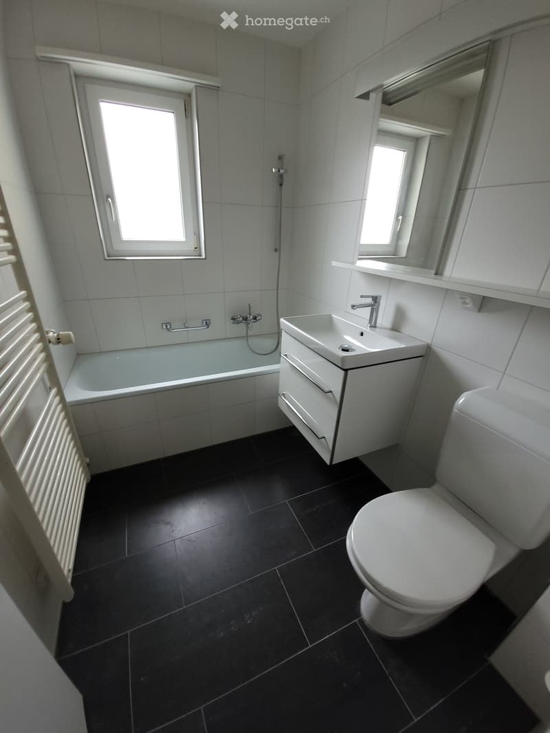 Bathroom. Modern bathroom with black tiled floor, white bathtub, sink, toilet, and mirror. Features two windows and a radiator on the left side.