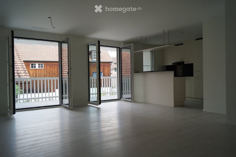 Empty room with wooden flooring, large sliding glass doors leading to a balcony, and a small kitchen area with cabinets visible in the background.