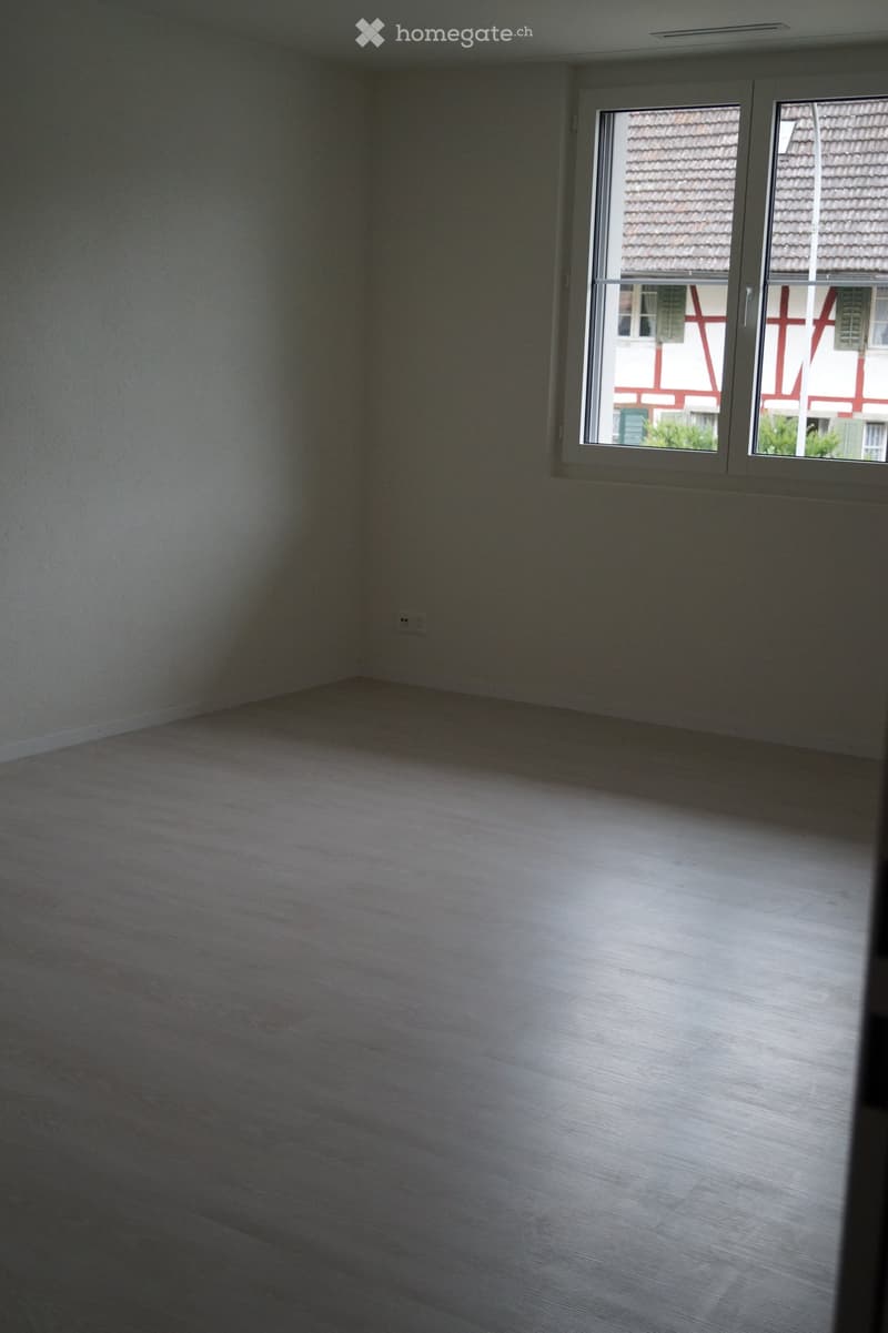 Empty room with light-colored wooden flooring and white walls, featuring a large window with a view of neighboring buildings.