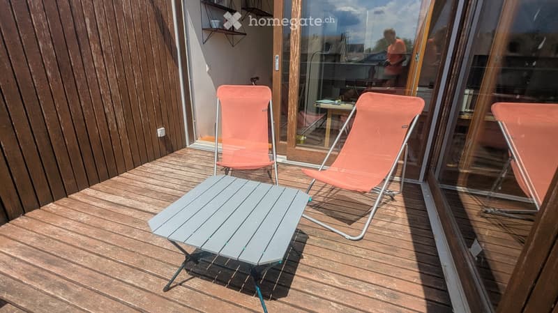Wooden deck with a grey folding table and three orange folding chairs, large glass doors leading inside.