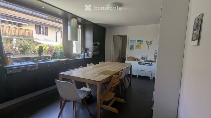 Modern kitchen with dark grey cabinets, a wooden dining table, white chairs, and various appliances including a dishwasher, sink, oven, and microwave. The space features large windows with a view of the balcony and outdoor area.