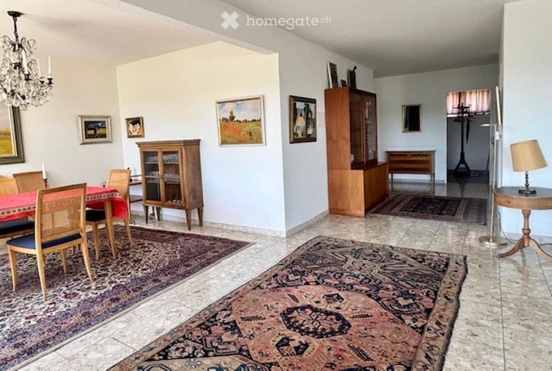 Dining area with a red tablecloth-covered dining table, wooden chairs, a wooden cabinet, side table with a lamp, and several picture frames on the walls.