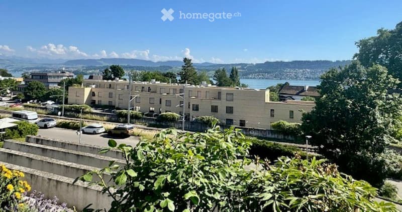 View of a multi-story residential building with parking lot, surrounded by greenery and a scenic view of a lake and mountains in the background.