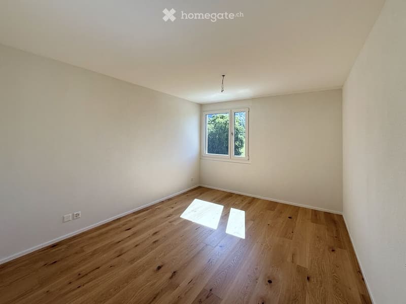 Empty room with light wooden flooring, white walls, and a single window showing greenery outside.